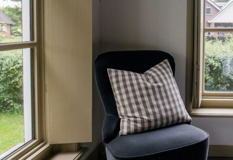 Close-up van een blauwe outlet stoel met een geruite kussen, naast een raam met uitzicht op een groene tuin. Grijs houten vloer in zacht licht.