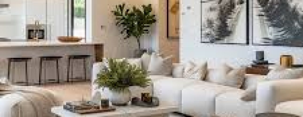 Een moderne woonkamer met een grote witte hoekbank, ronde voetenbankjes en een houten salontafel. Twee abstracte schilderijen hangen aan de muur. Op de achtergrond is er een keukenhoek met een aanrecht en barkrukken. Een grote potplant voegt groen toe.