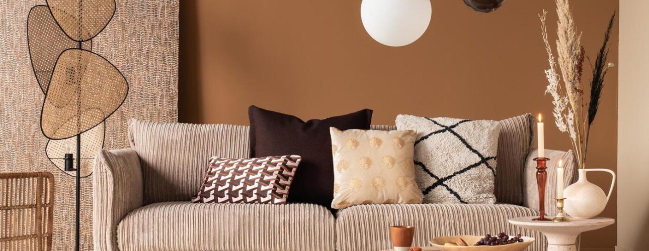 Een gezellige woonkamer met een beige getextureerde bank versierd met diverse kussens, een moderne lamp en een ronde houten salontafel. De kamer heeft natuurlijke tinten, een geweven staande lamp en een pluche wit tapijt op de houten vloer.