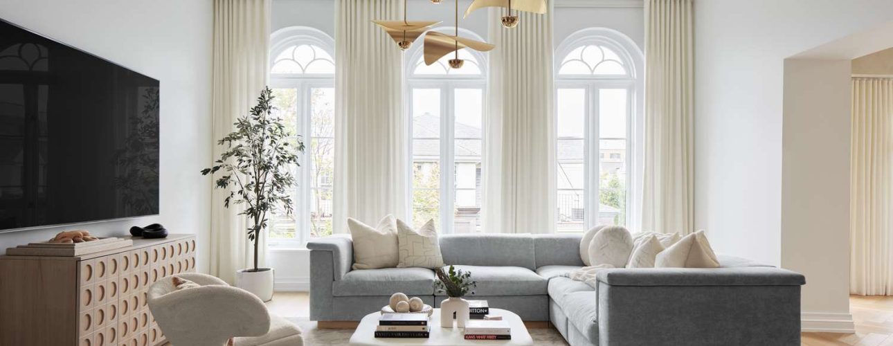 Een moderne woonkamer met een grote grijze sectionele bank, een crèmekleurige fauteuil, een unieke witte salontafel en een console met decoratieve items. Elegante hanglampen hangen aan het plafond en hoge ramen met gordijnen laten natuurlijk licht de ruimte vullen.