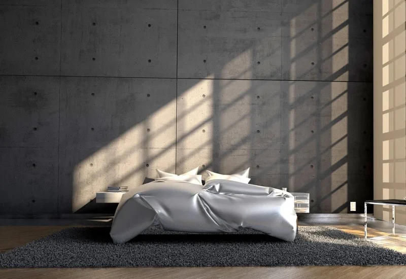 Minimalistische slaapkamer met betonnen muren en grote houten kozijnen die zonlicht binnenlaten. Een grijs vloerkleed en houten vloer.