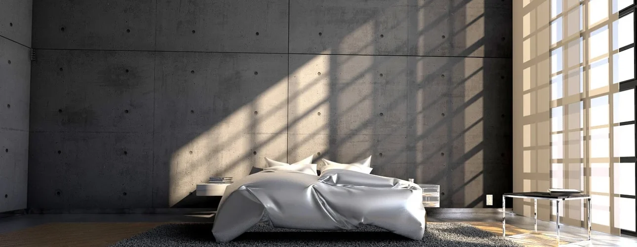 Minimalistische slaapkamer met betonnen muren en grote houten kozijnen die zonlicht binnenlaten. Een grijs vloerkleed en houten vloer.